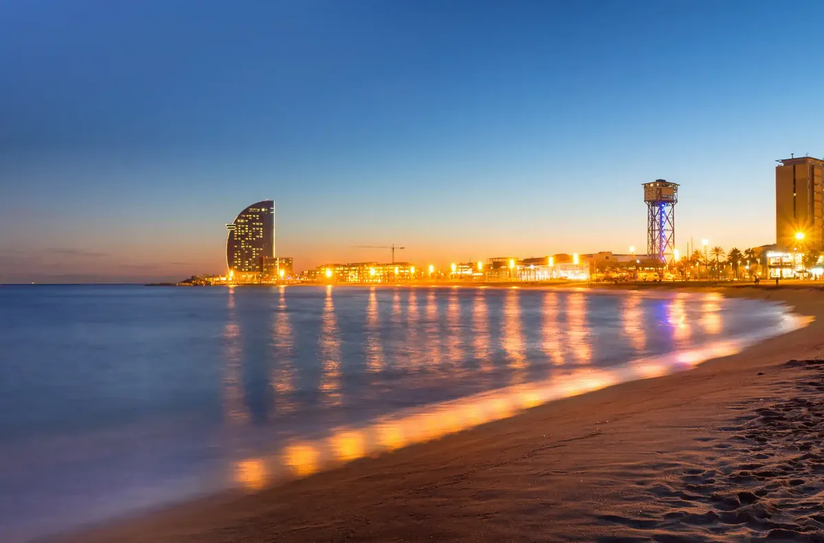 Der Strand Barceloneta in der Dämmerung, die Lichter der Strandpromenade spiegeln sich im ruhigen Meer