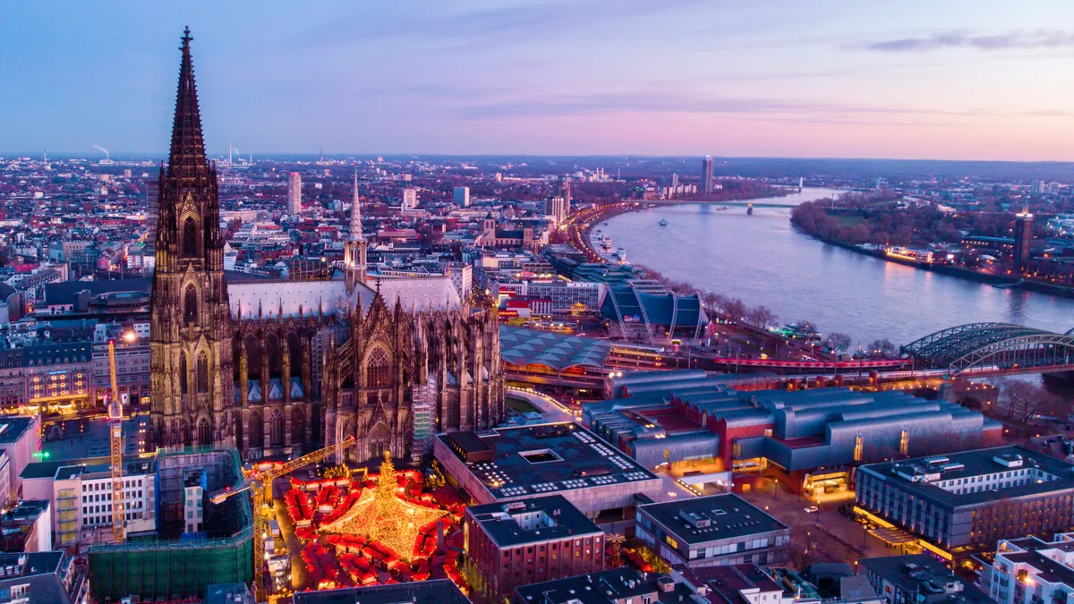 Der Kölner Dom in der Dämmerung von oben, an seinem Fuß leuchtet ein Weihnachtsmarkt, dahinter der Rhein