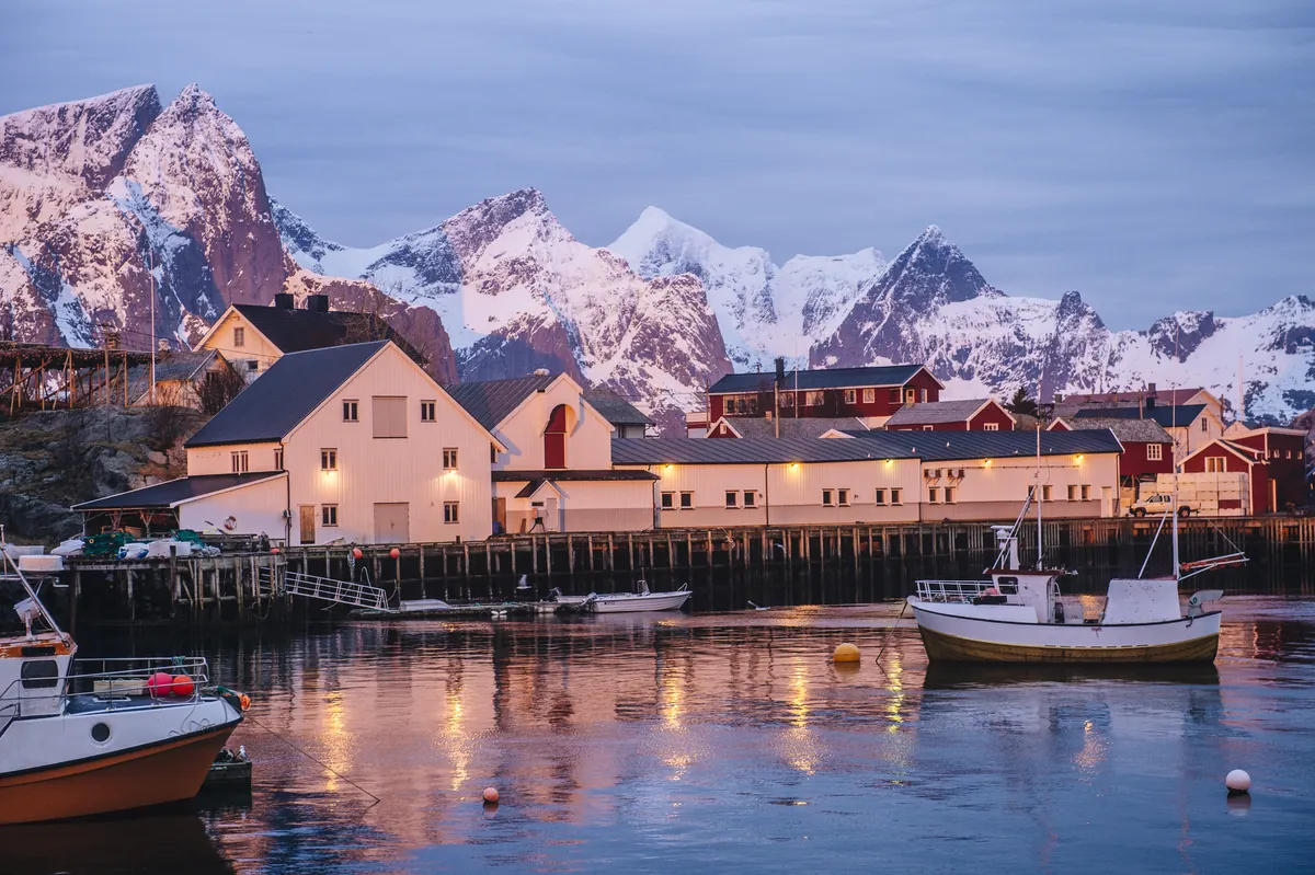 Der Hafen von Reine auf den Lofoten in der Dämmerung, beleuchtete Häuser und Boote unter schneebedeckten Gipfeln