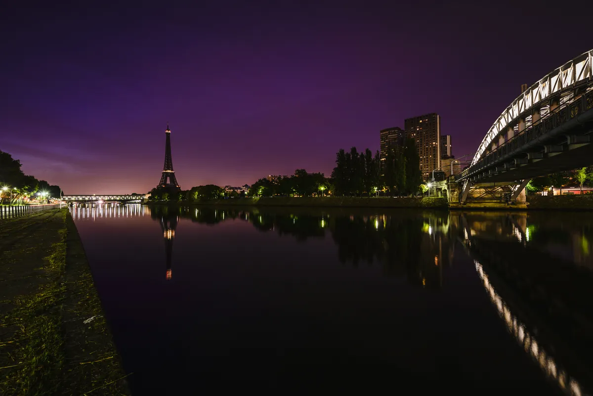 Die Seine in Paris in der Dämmerung unter violettem Himmel, eine beleuchtete Brücke spiegelt sich im stillen Wasser, am Horizont der Eiffelturm