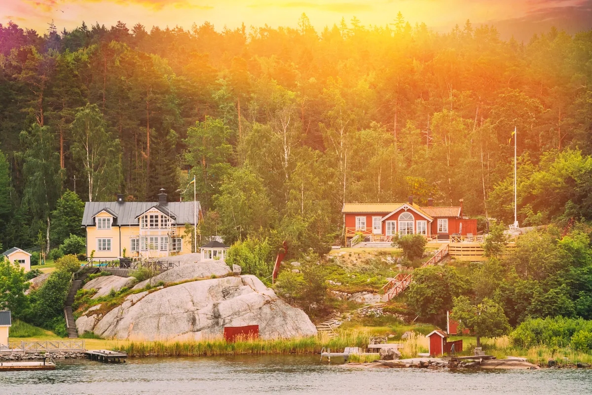 Ein rotes Holzhaus und eine gelbe Villa an einem felsigen Ufer an ruhigem Wasser, dahinter Kiefernwald im warmen Abendlicht
