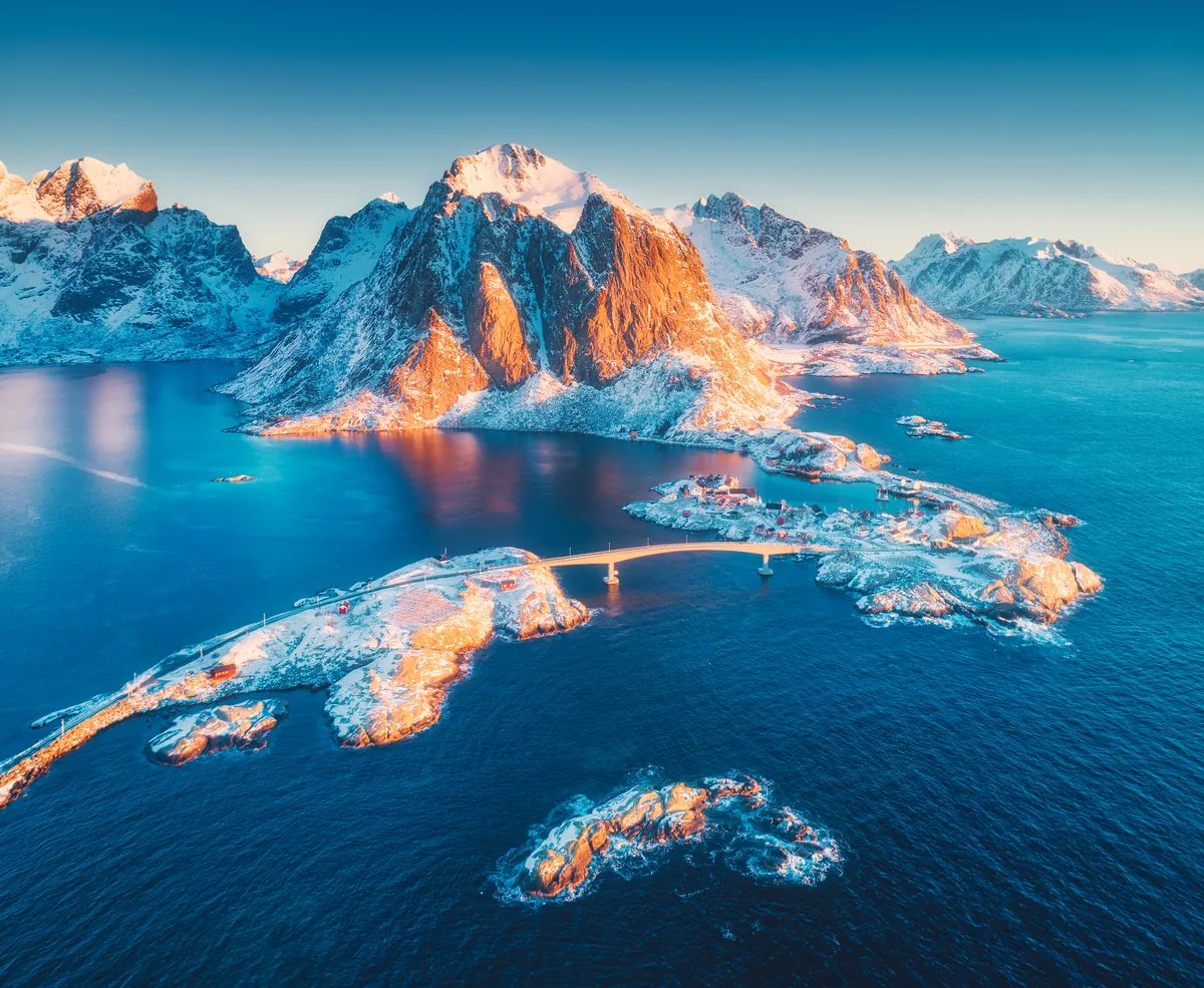 Luftbild der Lofotendörfer Reine und Hamnøy bei einem winterlichen Sonnenaufgang, die Gipfel leuchten über dem Meer