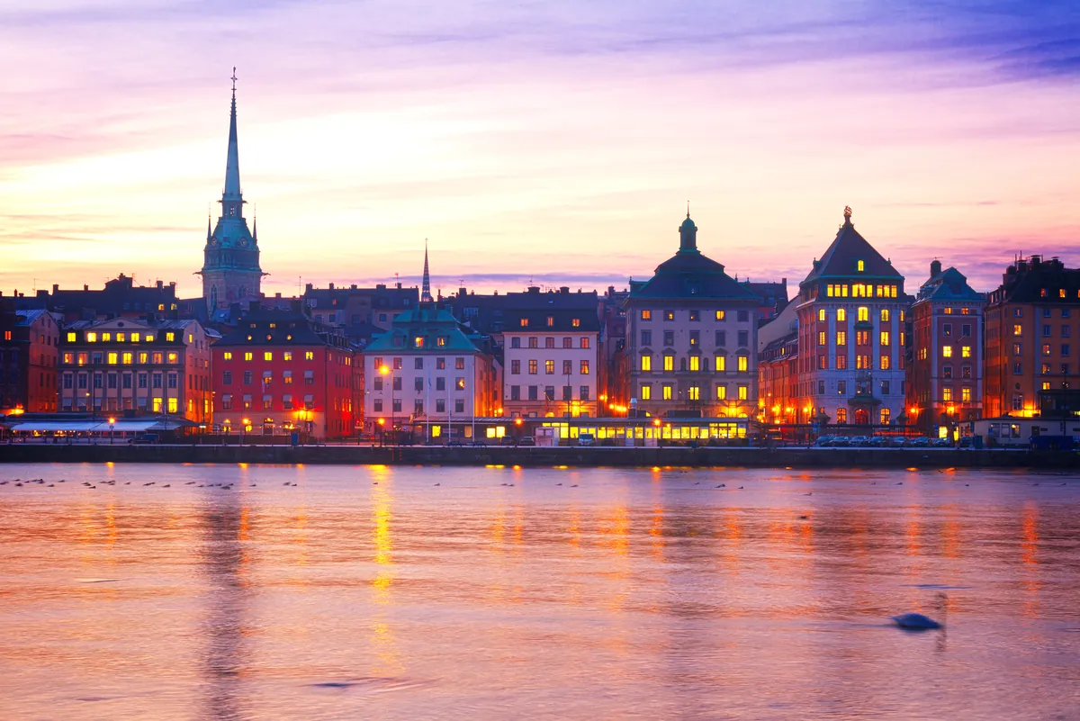 Die Altstadt von Stockholm in der Dämmerung unter rosa und violettem Himmel, Lichter spiegeln sich im Wasser